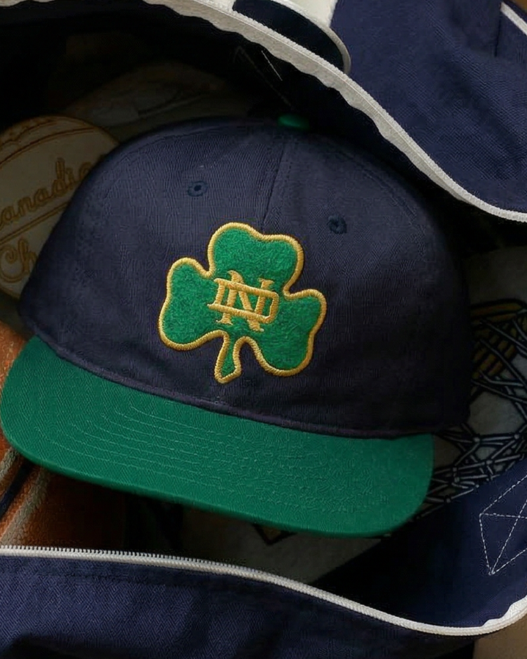 The Ebbets Field University of Notre Dame Shamrock Cotton Twill Ballcap features navy blue fabric, a green brim, and a green shamrock patch outlined in yellow with yellow ND letters on the front.