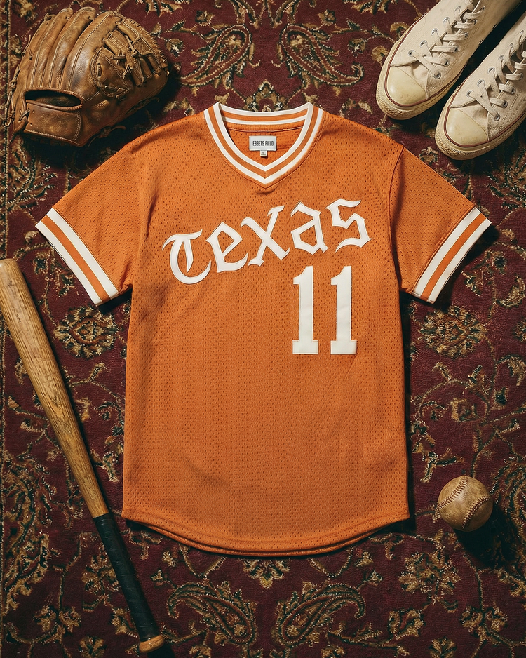 An orange University of Texas 1982 V-Neck Mesh Jersey by Ebbets Field, featuring the number 11, is displayed on a patterned rug with a baseball glove, bat, ball, and white sneakers nearby.