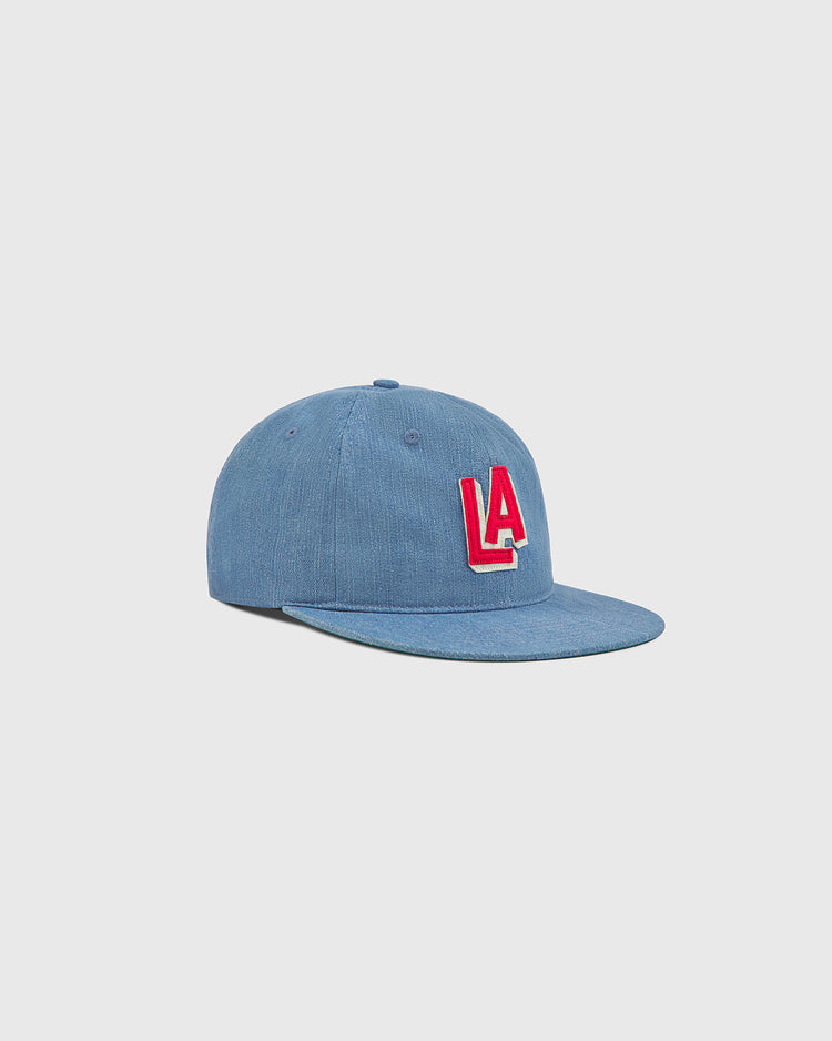 The Ebbets Field Los Angeles (PCL) Indigo Cotton Denim Ballcap features a flat brim and bold red LA lettering on the front, subtly evoking the classic style of a vintage Los Angeles Angels cap, shown against a simple light background.