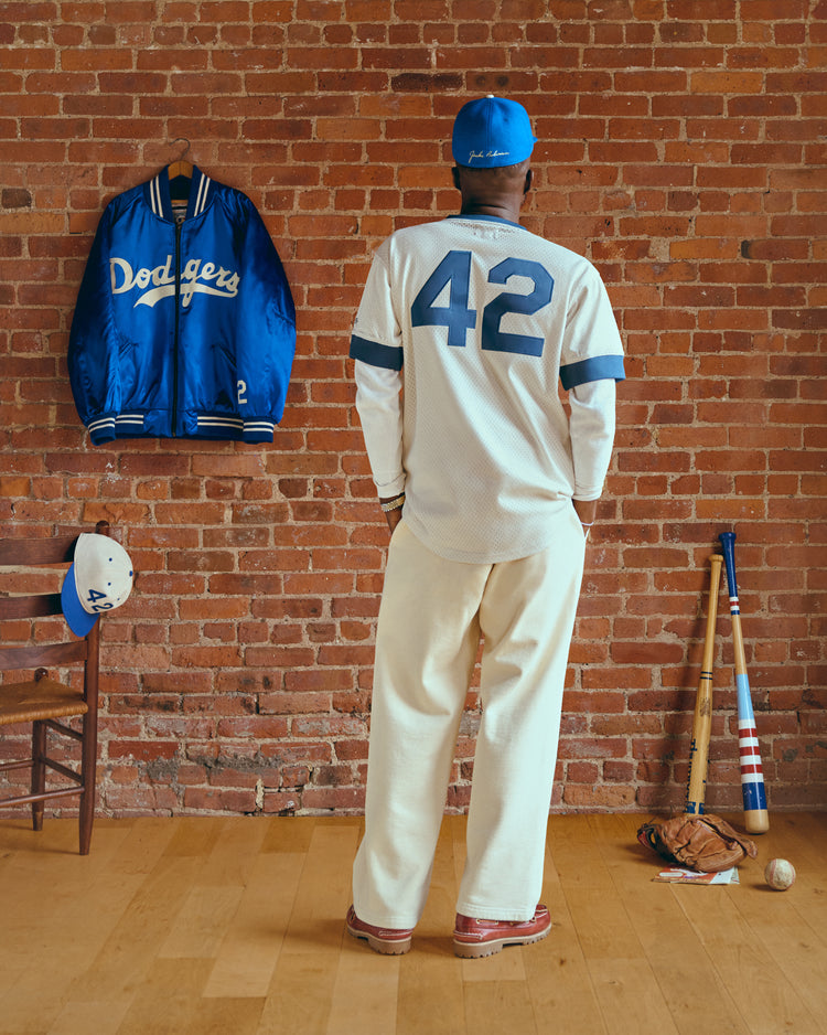 Wearing the Ebbets Field Jackie Robinson Mesh V-Neck Jersey and a blue cap, a fan stands by a brick wall with bats, glove, ball, Dodgers jacket, and cap nearby—capturing classic baseball spirit.