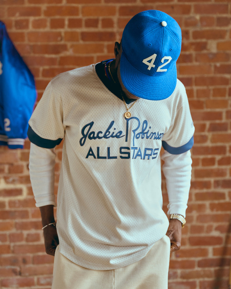 Against a brick wall, a person in an EFF Jackie Robinson Day Commemorative Wool Ballcap and a white Jackie Robinson All Stars jersey stands with head bowed, as a blue jacket hangs in the background.