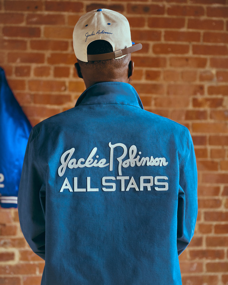 A person stands facing a brick wall, wearing an Ebbets Field Jackie Robinson Cotton Grounds Crew Jacket with ALL STARS on the back and a beige cap featuring blue embroidery.