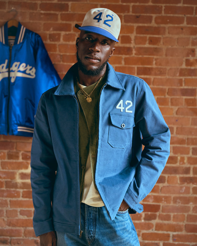 A man wearing a blue Ebbets Field Jackie Robinson Cotton Grounds Crew Jacket and matching cap, both featuring the number 42, stands before a brick wall with another Dodgers jacket hanging behind him.