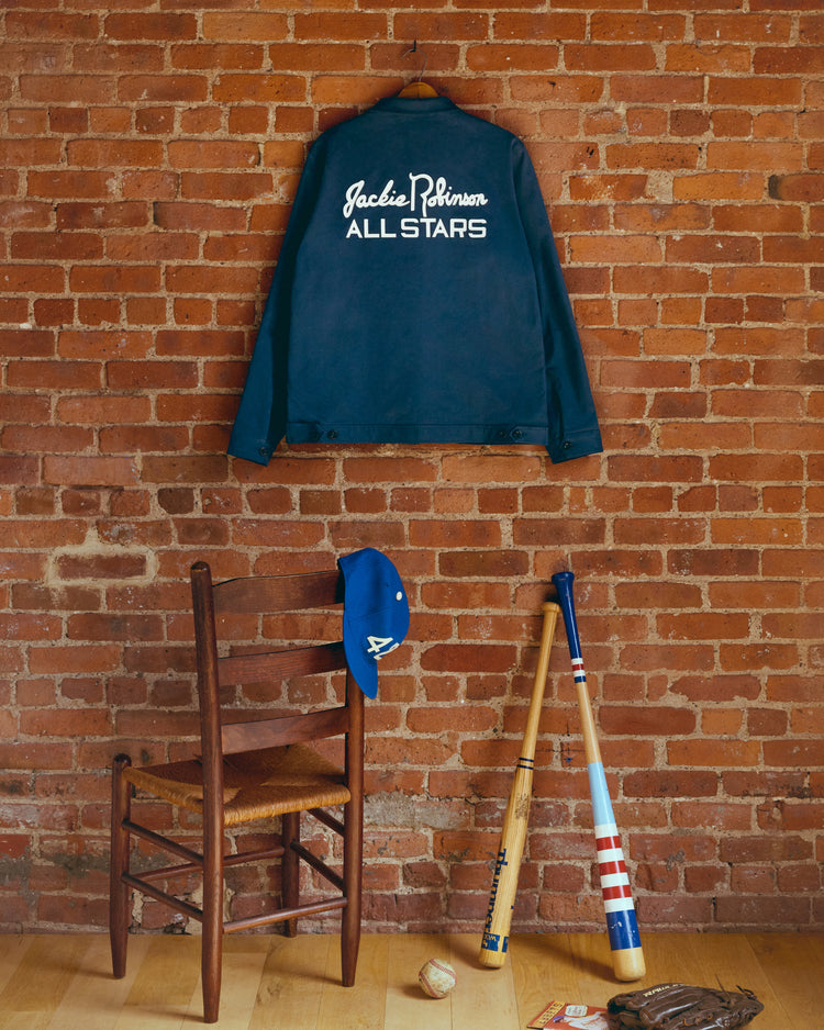 An Ebbets Field Jackie Robinson Cotton Grounds Crew Jacket in navy hangs on a brick wall above a wooden chair. A blue cap and glove rest on the chair, with two baseball bats and a baseball on the floor nearby.