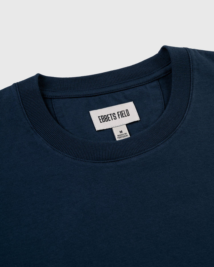 A close-up of the collar of the Ebbets Chest Logo Short Sleeve Tee by Ebbets Field Flannels, made from durable navy cotton. Two tags show the brand, size M (medium), and MADE IN KOREA—a comfortable, quality wardrobe staple.