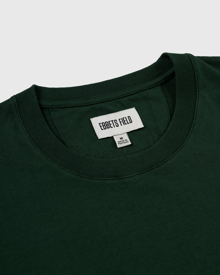 A close-up of the Ebbets Chest Logo Short Sleeve Tee by Ebbets Field Flannels, a durable dark green cotton crewneck with a white inner collar label reading EBBETS FIELD and M / PISTACHIO, set against a plain light background—an everyday staple.