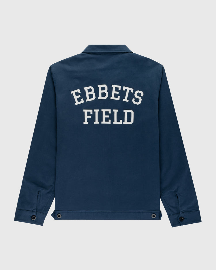 The Ebbets Field Cotton Grounds Crew Jacket by Ebbets Field Flannels is a navy cotton workwear jacket with long sleeves, a collar, buttoned cuffs, and EBBETS FIELD printed in bold white on the back—perfect for spring.