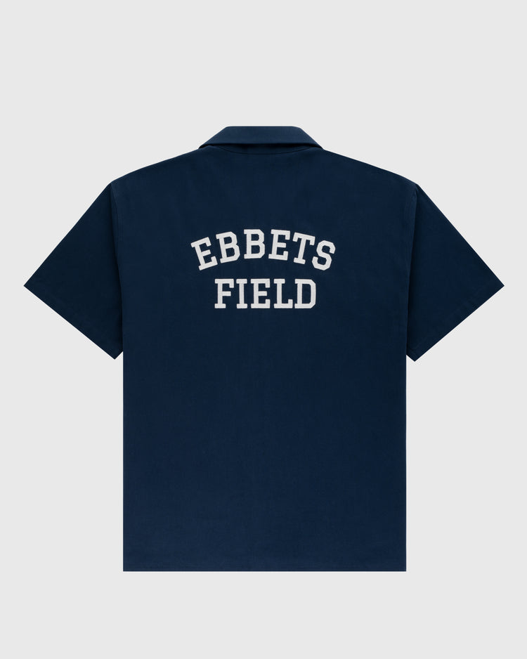 The Ebbets Field Flannels Ebbets Cotton Linen Grounds Crew Short Sleeve Workshirt in navy features “EBBETS FIELD” in large white letters on the back, shown against a plain light gray background.