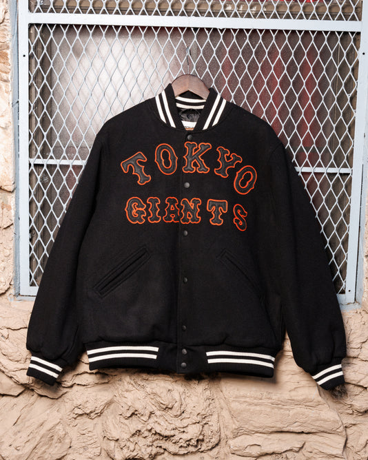 The Ebbets Field Tokyo Giants All-Wool Varsity Jacket, featuring bold orange lettering, hangs on a hanger against a wire mesh and stone wall backdrop, honoring Japanese baseball heritage.