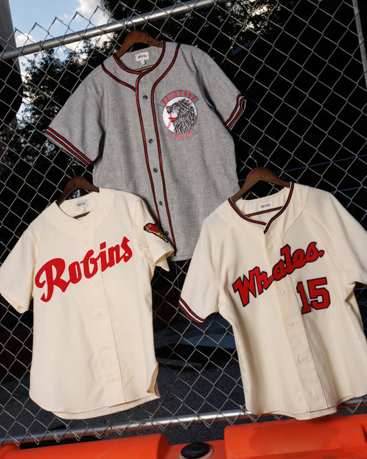 Three vintage-style baseball jerseys hang on a fence, including the Ebbets Field Shochiku Robins 1950 Authentic Wool Home Jersey, each featuring red and black accents on cream or gray fabric.