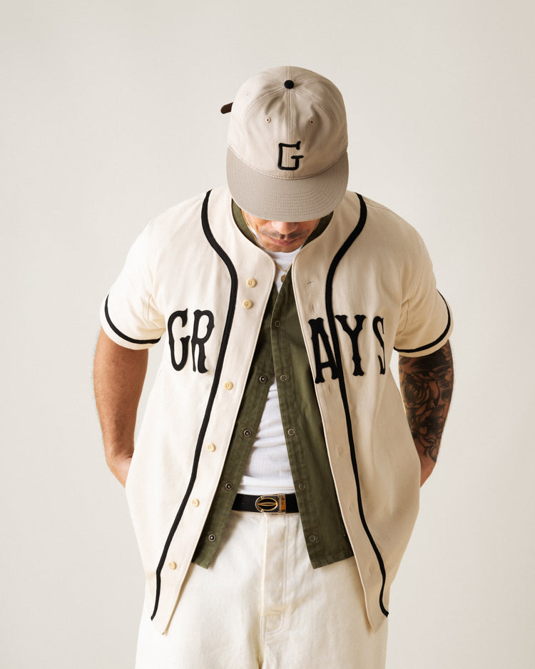 A person stands facing downward, arms behind their back, wearing a beige Ebbets Field Flannels Homestead Grays Cotton Twill Baseball Jersey inspired by the Negro Leagues, layered over an olive shirt with beige pants and a matching G-logo cap.