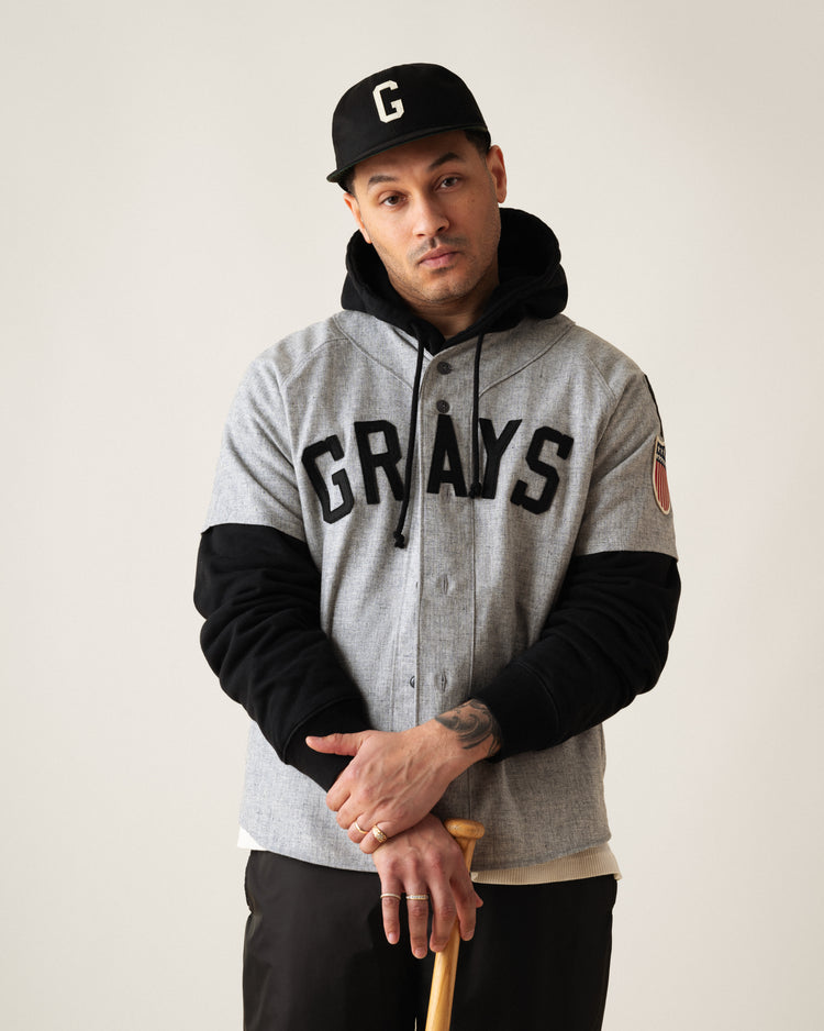 A man in a black cap, Ebbets Field Flannels Homestead Grays 1944 Authentic Wool Road Baseball Jersey over a black hoodie, and black pants holds a bat and looks at the camera with a neutral expression.