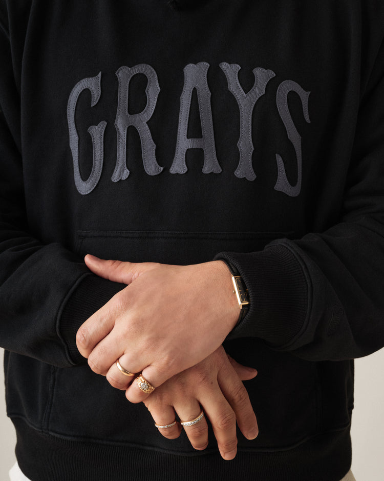 A person in an Ebbets Field Flannels Homestead Grays Heavyweight Cotton Terry Pullover Hoodie, featuring bold gray GRAYS lettering, stands with crossed hands, showing rings and a watch—celebrating Negro Leagues Baseball Museum heritage.