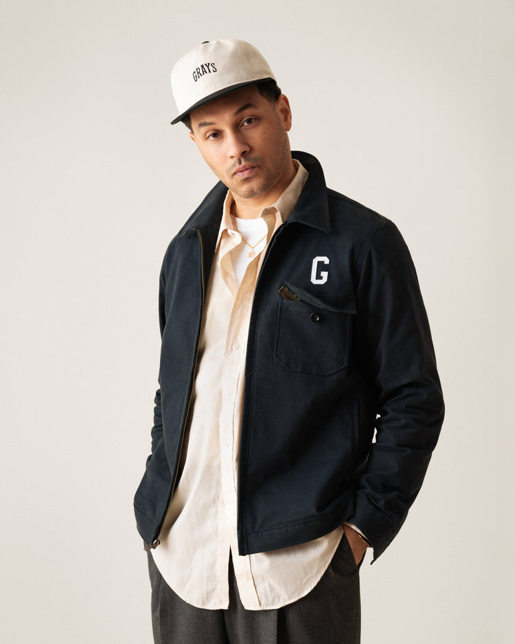 A man stands with hands in pockets against a plain background, wearing an Ebbets Field Flannels Homestead Grays Embroidered Two Tone Cotton Twill Ballcap, a black jacket with a G on the chest, cream shirt, and gray pants.
