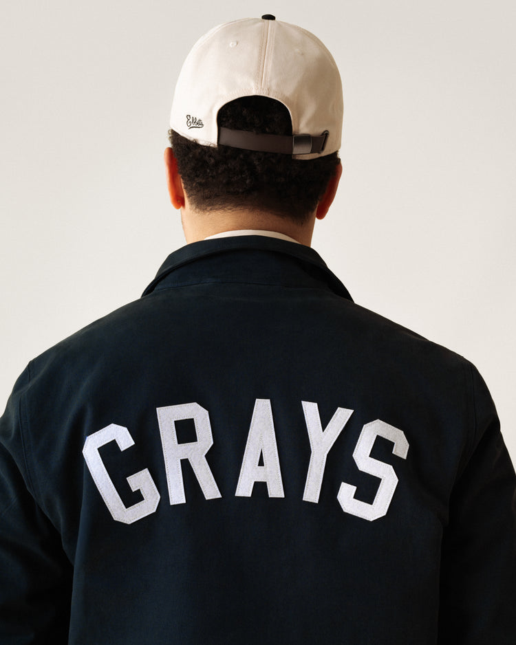 A person is shown from behind in a black Ebbets Field Flannels Homestead Grays Cotton Grounds Crew Jacket with “GRAYS” in large white letters on the back and a white baseball cap, against a plain background.