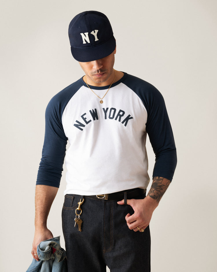 A man in an Ebbets Field New York Black Yankees Felt NY Cotton Twill Ballcap and a white raglan shirt holds a denim jacket and keys, one hand in his pocket, standing against a plain background.