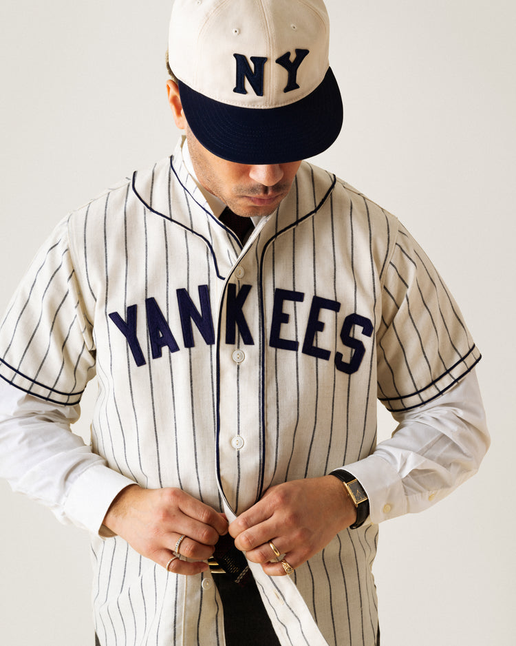 A person wearing an Ebbets Field New York Black Yankees 1935 Authentic Wool Home Baseball Jersey and an NY cap looks down while buttoning the jersey, styled with a white shirt, wristwatch, and several rings.