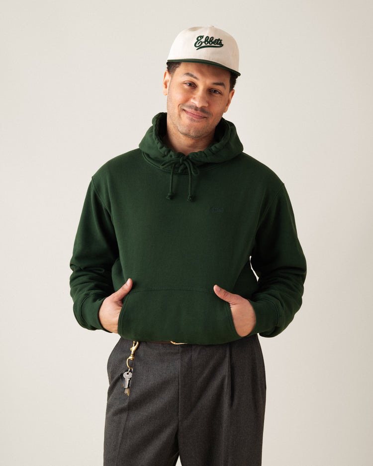 A man smiles with his hands in the pocket of an Ebbets Field Flannels Ebbets Chest Logo Heavyweight Cotton Terry Pullover Hoodie in dark green, paired with gray pants and a white cap with green lettering; keys hang from his belt loop.