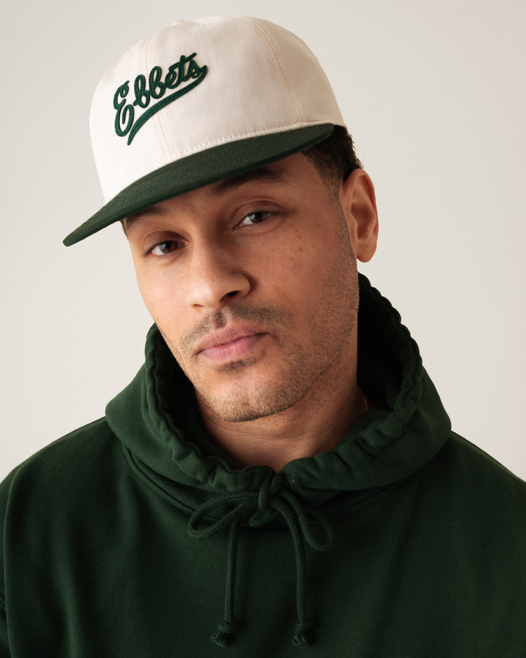 A man in a cream and green Ebbets Field Flannels Ebbets Felt Script Cotton Twill Ballcap and a forest green hoodie looks at the camera with a neutral expression, set against a plain, light-colored background.
