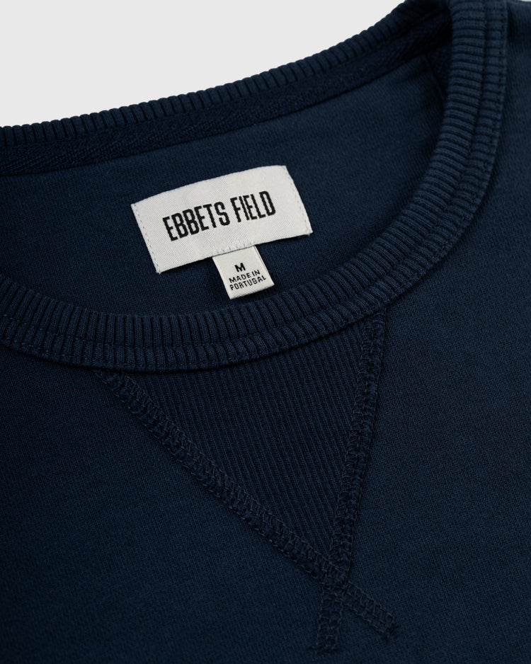 Close-up of the Ebbets Field Flannels Ebbets Chest Logo Heavyweight Cotton Terry Crewneck in navy, featuring a ribbed collar, V-stitch at the neckline, and white EBBETS FIELD label with an M Made in Portugal tag.