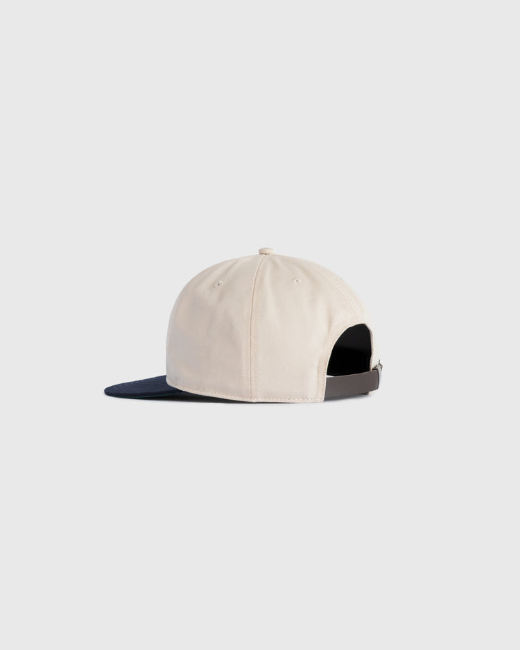 The Ebbets Field Flannels Felt Script Cotton Twill Ballcap, shown in beige with a dark navy brim, features a black adjustable strap and silver buckle. Crafted from cotton twill, this cap blends classic style and comfort against a light background.