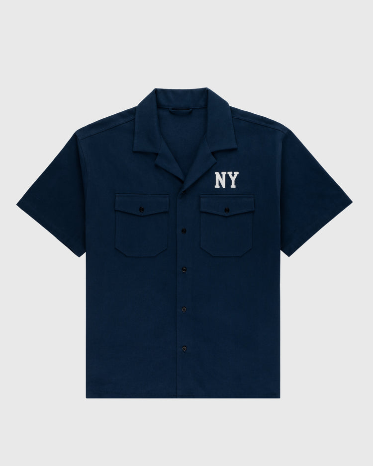 The Ebbets Field Flannels Cotton Linen Grounds Crew Short Sleeve Workshirt in navy blue features two button-flap chest pockets and NY in white above the left pocket. Shirt is displayed flat on a light gray background.