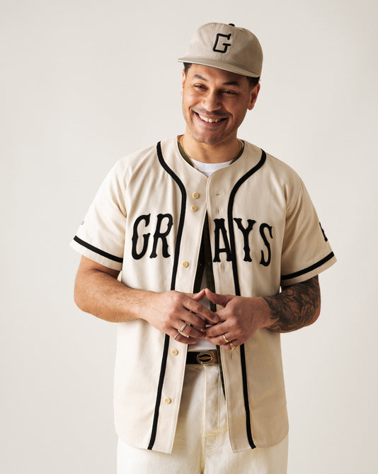 A smiling man wearing a cream-colored baseball jersey with GRAYS on the front, a matching cap with a G, white pants, and gold jewelry stands against a plain background.