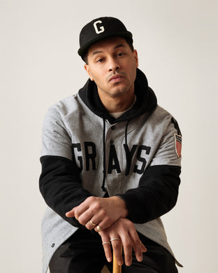 A man wearing a black and grey Grays hoodie and a black cap with a G sits on a stool, leaning forward with his hands resting in front, looking straight at the camera with a neutral expression.