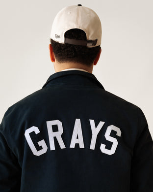 A person wearing a two-tone baseball cap and a dark jacket with the word GRAYS in large white letters on the back is shown from behind against a plain background.