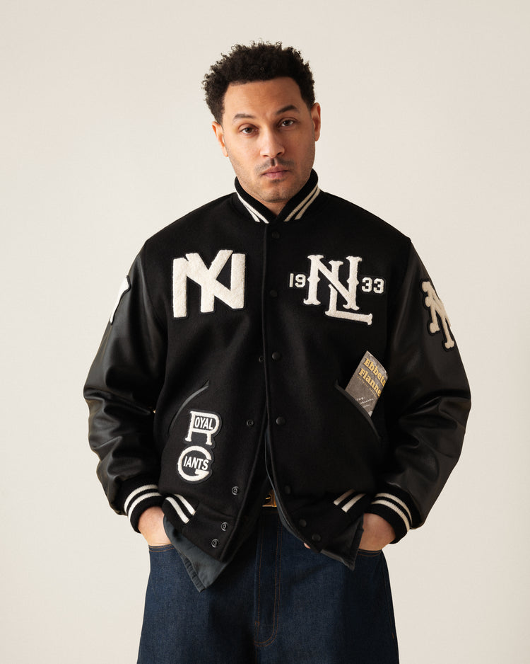 A man models the Ebbets Field Negro League Wool & Leather Varsity Jacket, featuring a black body, white NY and 1933 patches, leather sleeves, and striped cuffs, standing with hands in pockets against a plain light background.