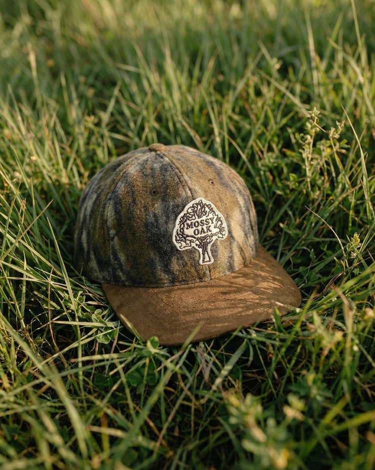 The Ebbets Field Mossy Oak x Vintage Tree Logo Ballcap, featuring Bottomland camo and a brown brim, sits on green grass among tall blades and small plants in natural sunlight.
