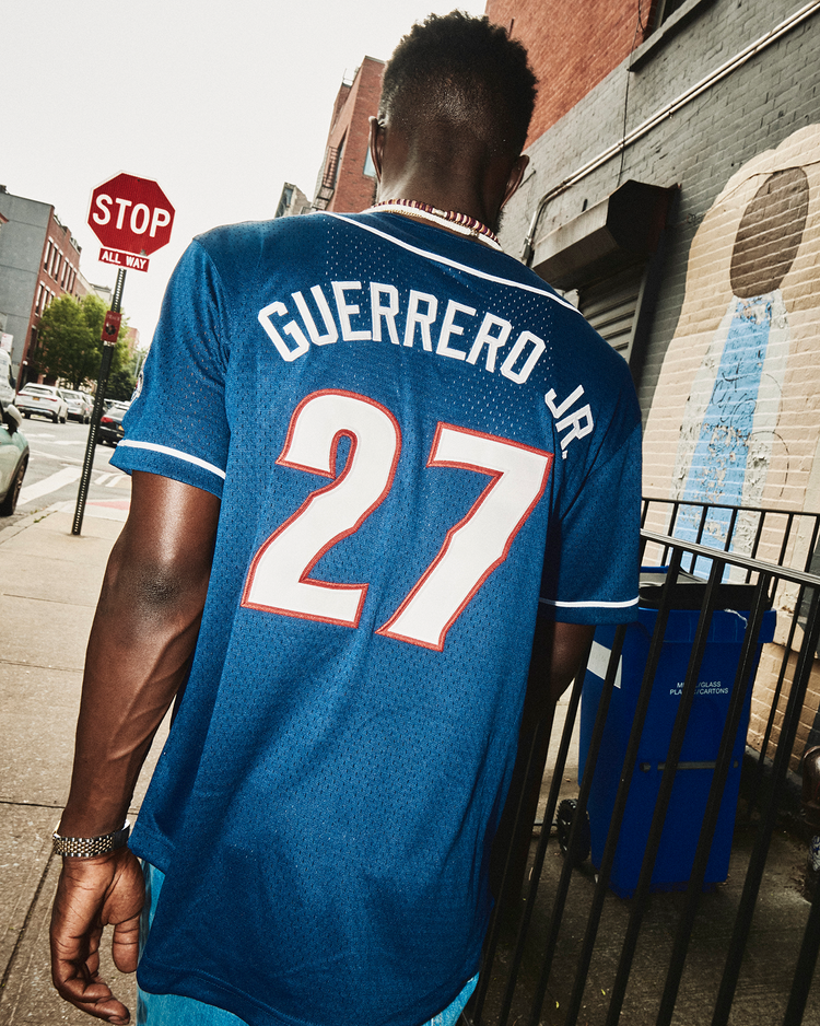 A man wearing an Ebbets Field Vladimir Guerrero Jr. New Hampshire Fisher Cats Mesh Button-Up Jersey walks down a city sidewalk past a stop sign and a mural on a brick wall, sporting this throwback minor league look.