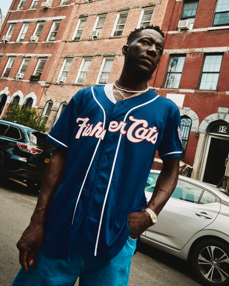 A man wearing an Ebbets Field Vladimir Guerrero Jr. New Hampshire Fisher Cats Mesh Button-Up Jersey and blue jeans stands on a city street in front of a red brick building, looking to the side with one hand in his pocket.