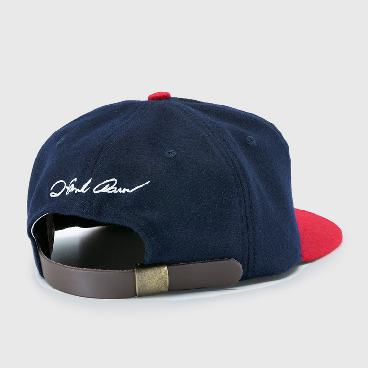 The Ebbets Field Flannels Hank Aaron 1957 Signature Series Ballcap features navy wool, a red brim, a brown leather strap with brass clasp, white cursive embroidery near the back opening, and is proudly made in the USA.