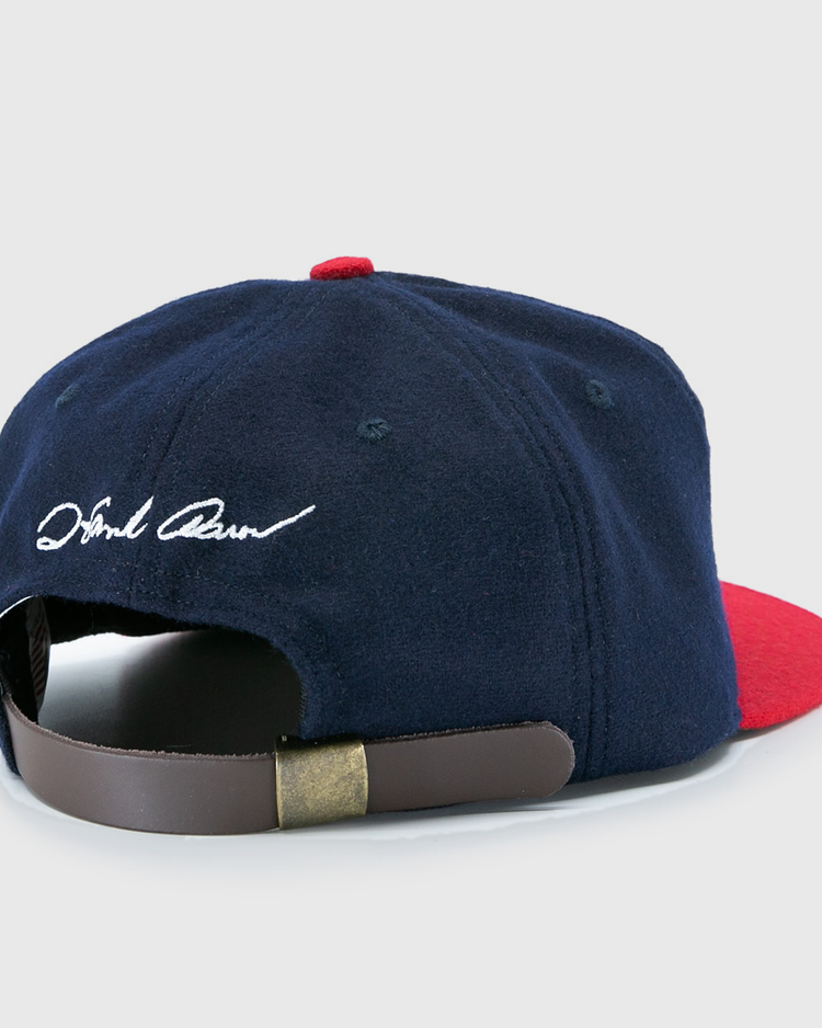 The Ebbets Field Flannels Hank Aaron 1957 Signature Series Ballcap features navy wool, a red brim, a brown leather strap with brass clasp, white cursive embroidery near the back opening, and is proudly made in the USA.