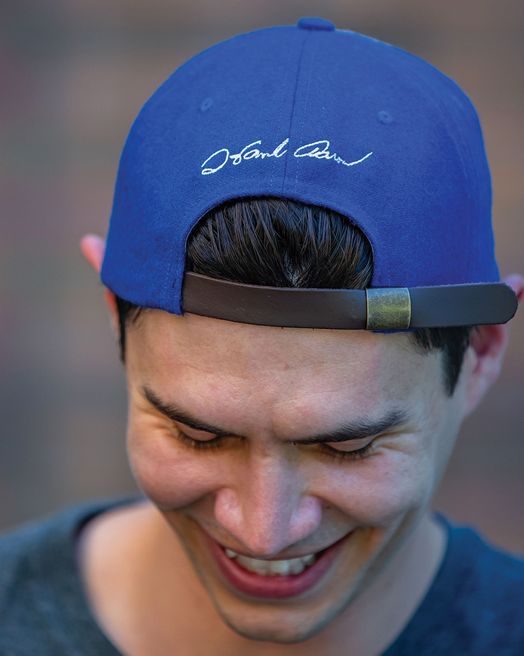 A smiling person wears the Ebbets Field Flannels Hank Aaron 1973 Signature Series Ballcap backwards, revealing white cursive writing on the back as they look down, with a blurred background behind them.