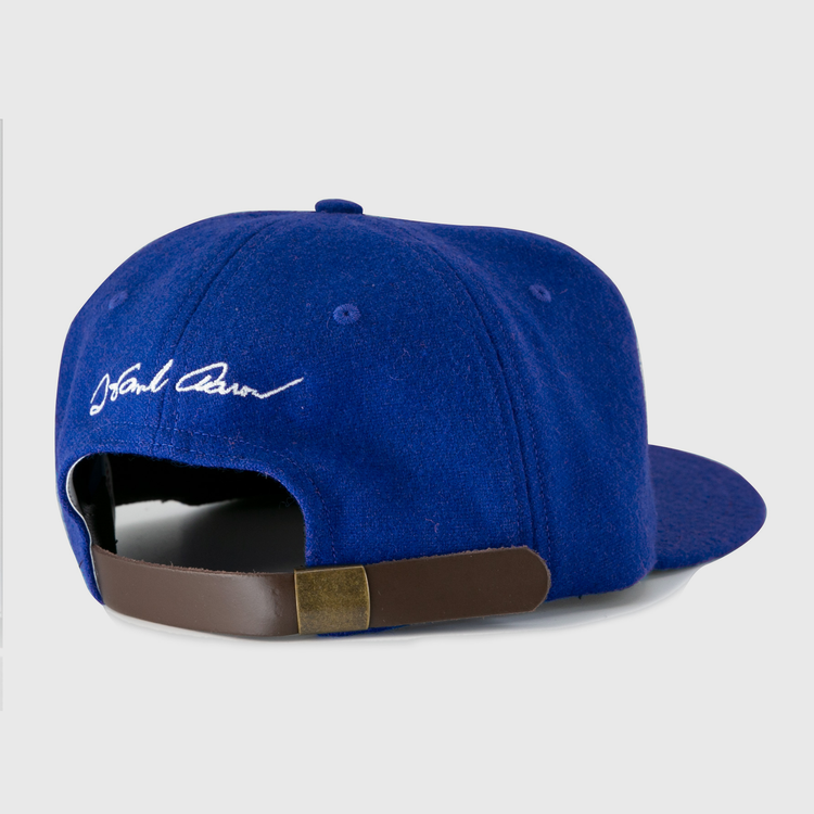 The Ebbets Field Flannels Hank Aaron 1973 Signature Series Ballcap features a royal blue wool design, white embroidered signature above an adjustable brown leather strap with brass buckle, and is proudly made in the USA.