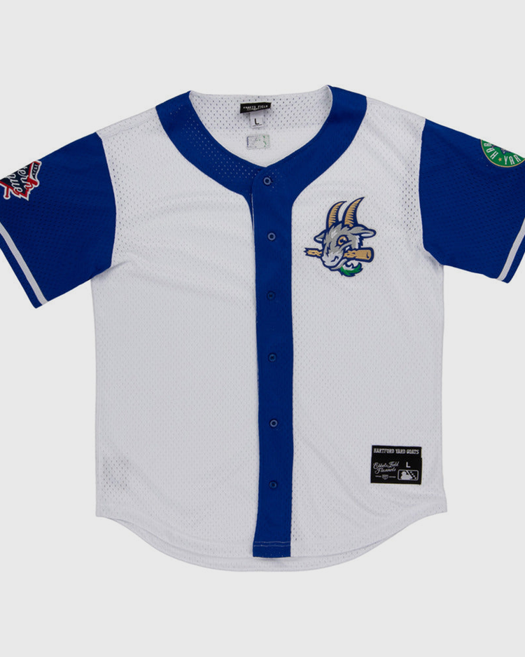 The Ebbets Field Hartford Yard Goats MiLB Button Down Jersey is white with blue sleeves and trim, a cartoon goat logo on the chest, sleeve patches, button-down front, and a black label near the hem.