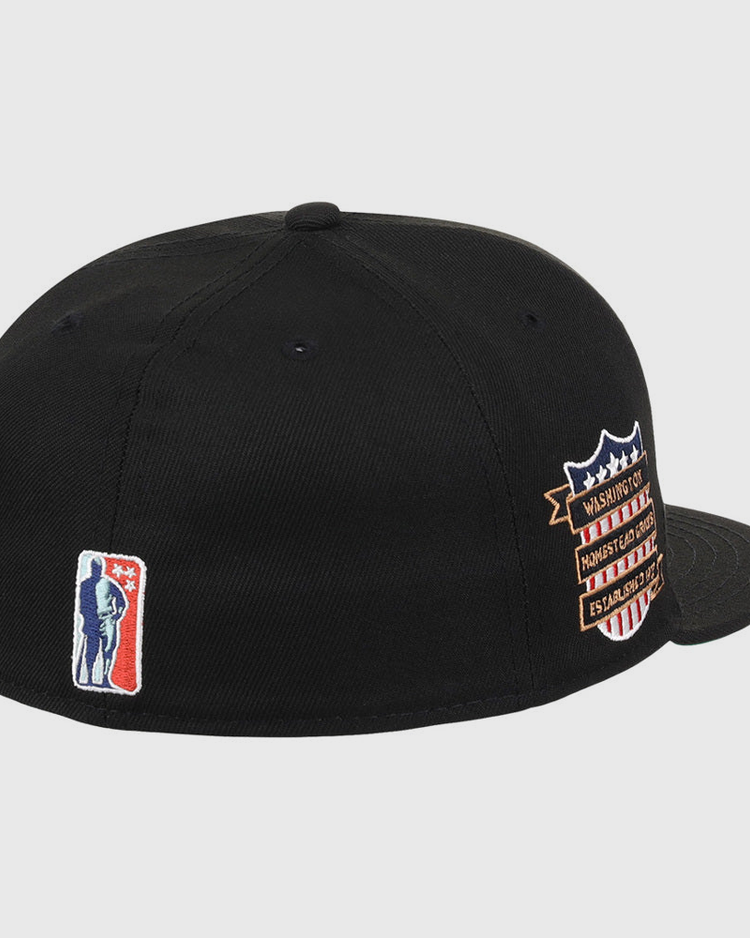 The Ebbets Field Homestead Grays NLB Tc Fitted Ballcap is a black cap featuring a small blue and white basketball player silhouette embroidered on the back and a multicolored shield patch with text on the side.