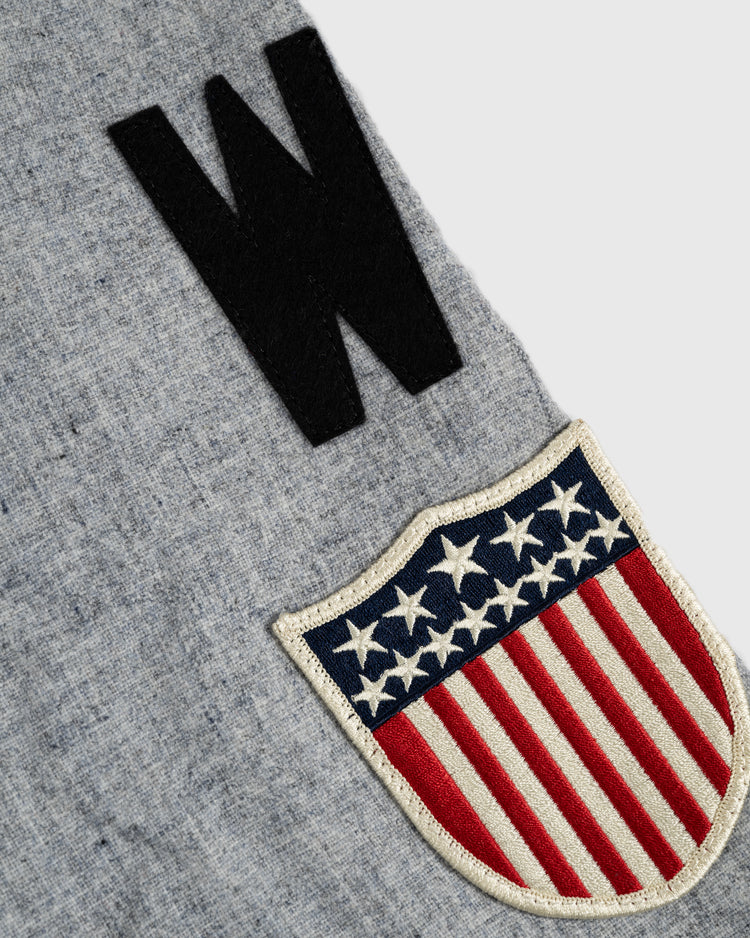 A close-up of the Ebbets Field Flannels Homestead Grays 1944 Authentic Wool Road Baseball Jersey shows gray wool with a large black W and an embroidered shield patch with stars and red stripes, evoking the American flag.