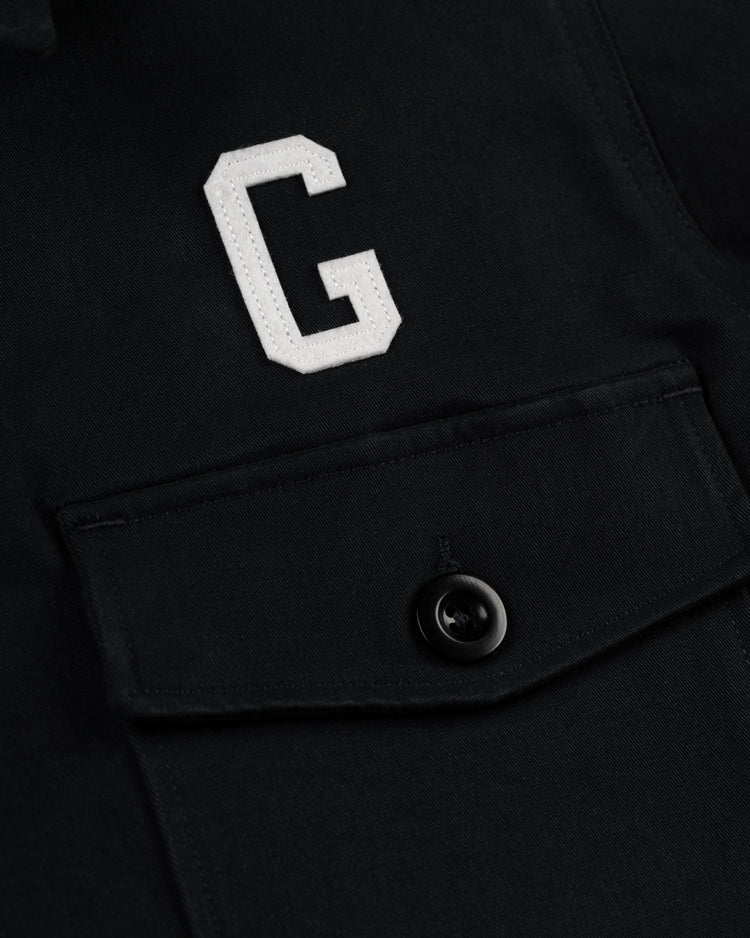 Close-up of the Ebbets Field Flannels Homestead Grays Cotton Grounds Crew Jacket—a black cotton twill piece with a flap pocket, black button, and a large white stitched G above the pocket, honoring the legendary Negro Leagues team.