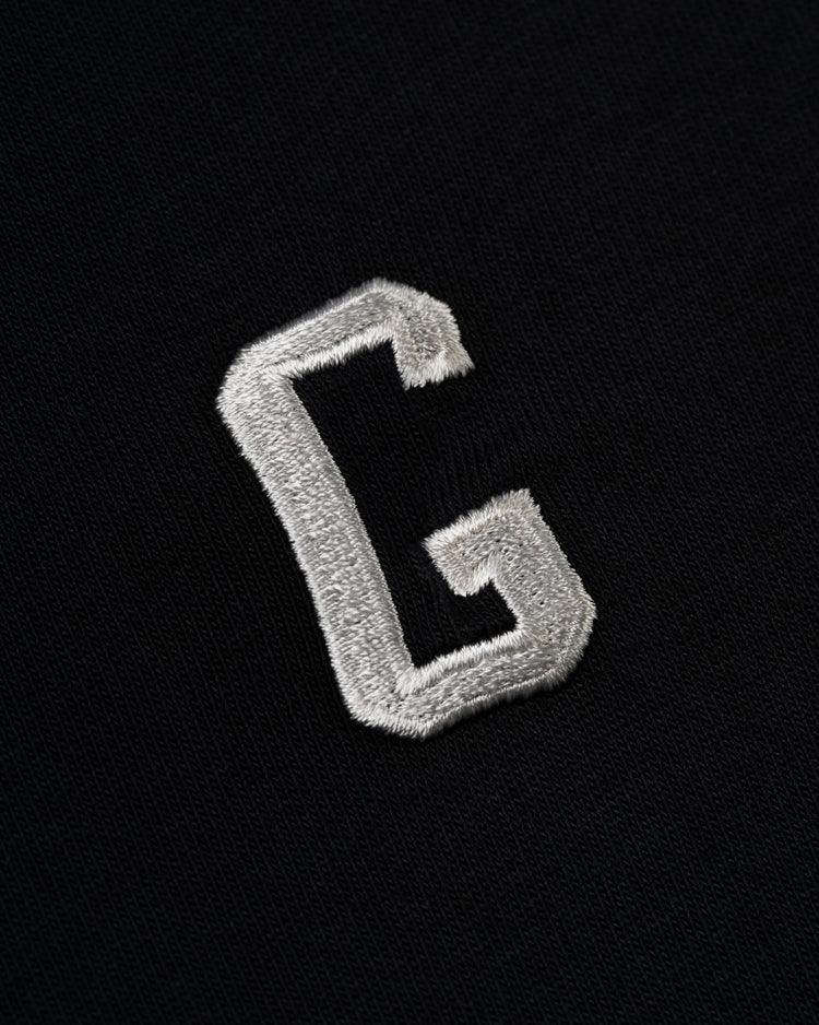A close-up of the white, embroidered G on black fabric from the Homestead Grays Embroidered G Short Sleeve Tee by Ebbets Field Flannels, honoring Josh Gibson and the Homestead Grays at the Negro Leagues Baseball Museum.