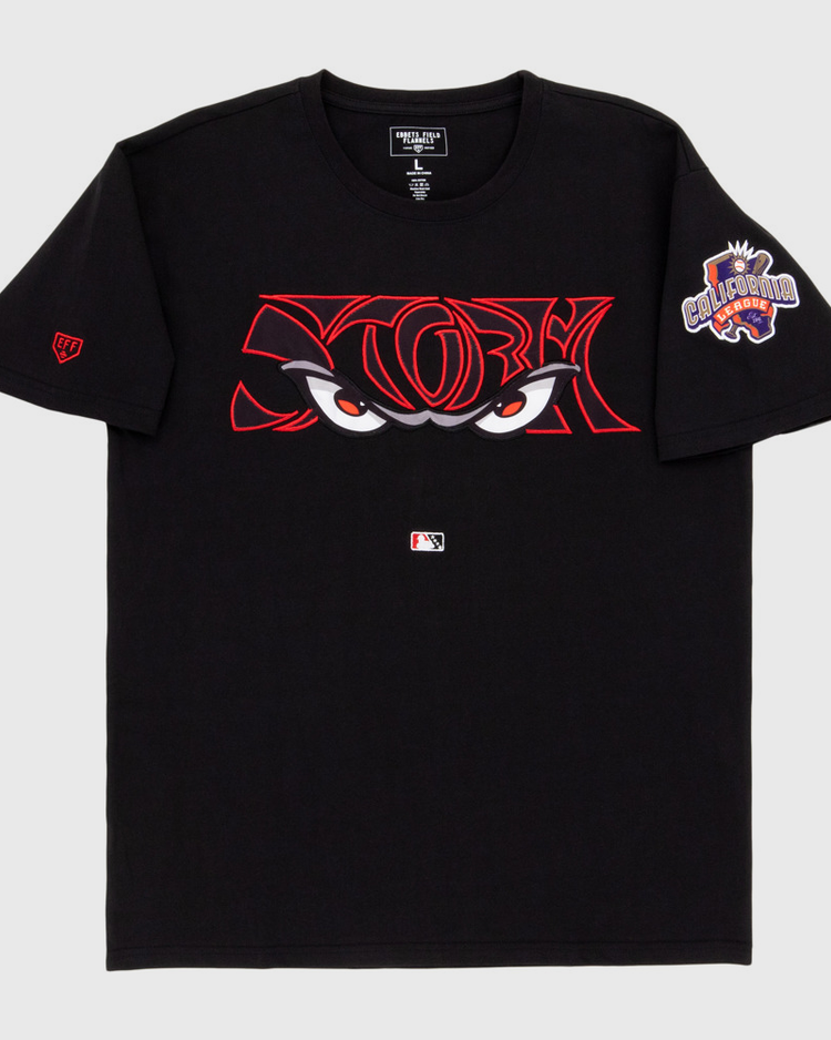 The Ebbets Field Lake Elsinore Storm MiLB Fashion Cotton Short Sleeve Tee is a black shirt with bold red-outlined cartoon eyes on the chest, an MLB logo underneath, and a colorful 