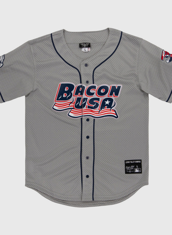 The Lehigh Valley Ironpigs MiLB Button Down Jersey by Ebbets Field is a gray baseball jersey with navy trim, “Bacon USA” in red, white, and blue on the front, plus cartoon bacon designs on the left sleeve and right shoulder.