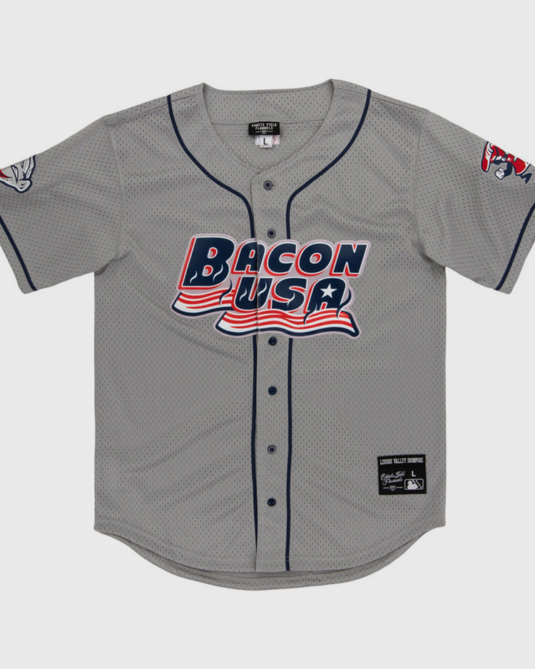 The Lehigh Valley Ironpigs MiLB Button Down Jersey by Ebbets Field is a gray baseball jersey with navy trim, “Bacon USA” in red, white, and blue on the front, plus cartoon bacon designs on the left sleeve and right shoulder.