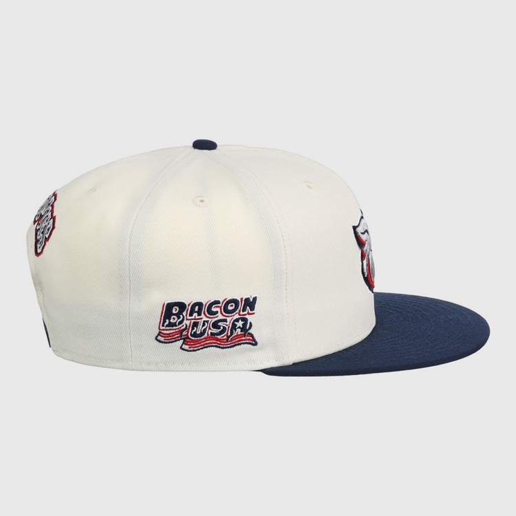 The Lehigh Valley Ironpigs MiLB Snapback Ballcap by Ebbets Field is a white cap with a navy brim, embroidered Bacon USA text and flag on the side, plus additional patches on the front and back.