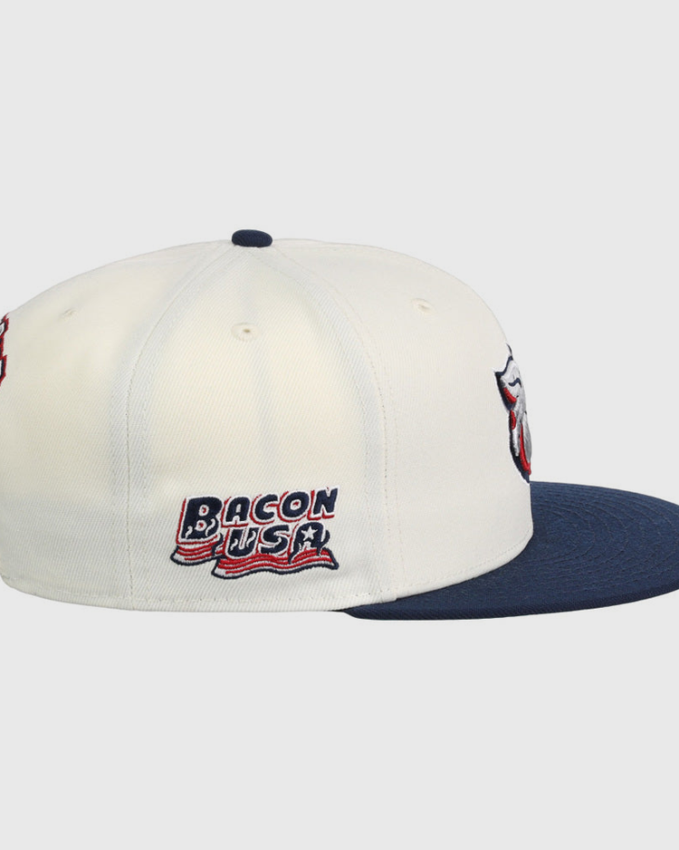 The Lehigh Valley Ironpigs MiLB Snapback Ballcap by Ebbets Field is a white cap with a navy brim, embroidered Bacon USA text and flag on the side, plus additional patches on the front and back.