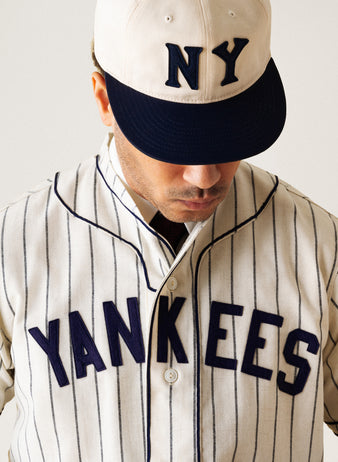 A person wearing a cream pinstripe baseball jersey with YANKEES in navy lettering and a matching NY cap looks downward, obscuring their eyes. The background is plain and light-colored.