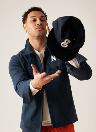 A man in a navy jacket tosses a black baseball cap with a white logo into the air. He is looking up at the hat with a neutral expression against a plain light background.