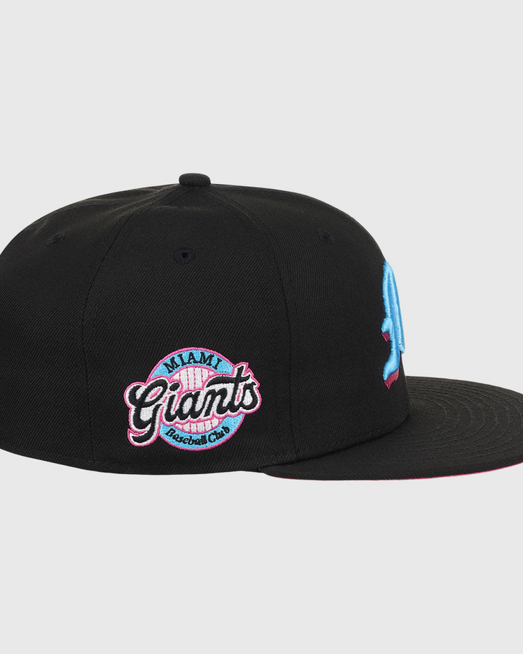 The EFF Miami Giants NLB Team Color Fitted Ballcap is a vintage-inspired black cap with an embroidered light blue M on the front and a pink, blue, and white circular Miami Giants logo on the side, honoring a classic Black barnstorming team.